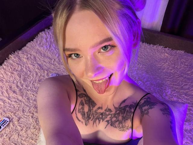 EnchantingEyes with blonde hair, green eyes, and tattoos on chest and arms, sticking out tongue, wearing black top, on purple-lit white fluffy rug, remote control on left.