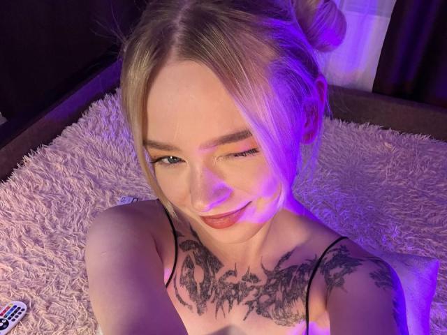 EnchantingEyes with blonde hair in buns, winking, smiling, showing tattoos on chest, wearing a black spaghetti strap top, on a pink shaggy rug, with purple lighting.