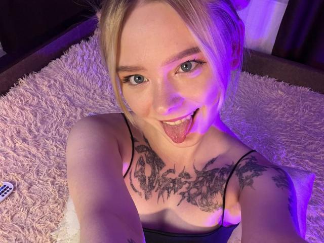 EnchantingEyes with blonde hair, blue eyes, and tattoos on her chest and shoulder, sticking out her tongue, wearing a black spaghetti strap top, on a pink shaggy rug.