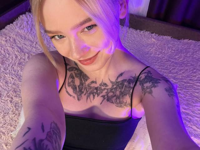EnchantingEyes with blonde hair, tattoos on chest and arm, smiling, wearing black top, purple lighting, lying on fluffy white rug, taking selfie, showing tits, cute, sexy.