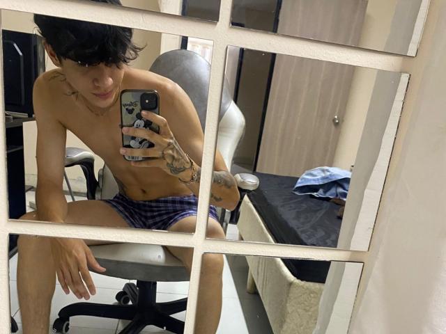 Mirror selfie of KataAndByron, shirtless with tattoos, wearing plaid underwear, sitting on a chair, holding a phone with a black and white case, in a room with a bed.