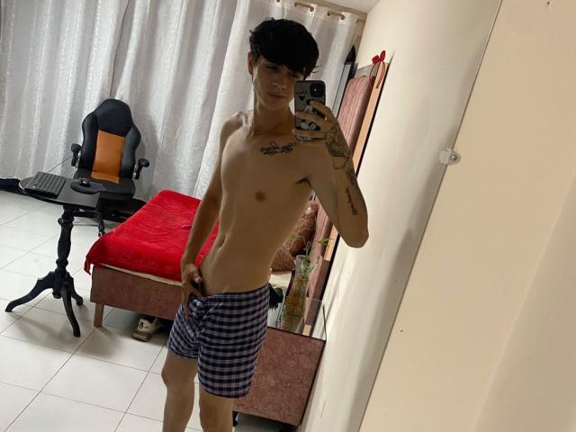 Shirtless KataAndByron with tattoos, wearing blue checkered shorts, holding phone, standing in bedroom, mirror selfie, black hair, slim build, small ass, visible penis bulge, white tiled floor.
