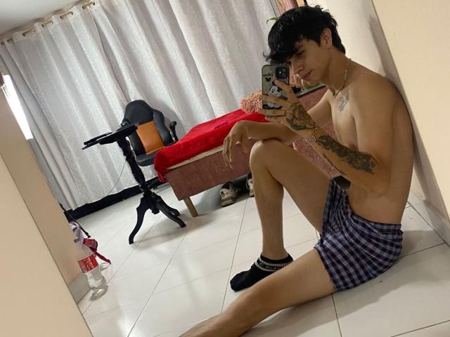KataAndByron, shirtless, tattooed, in checkered shorts, black socks, sitting on tile floor, taking selfie in mirror, red bed in background, white curtains, holding phone, curly black hair.