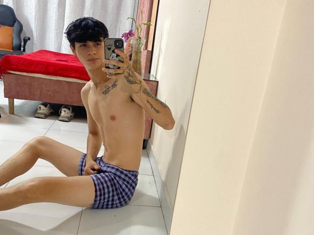 KataAndByron, shirtless with black curly hair, tattoos on chest and arm, wearing purple checkered shorts, sitting on white tiled floor, taking mirror selfie, holding phone in right hand.