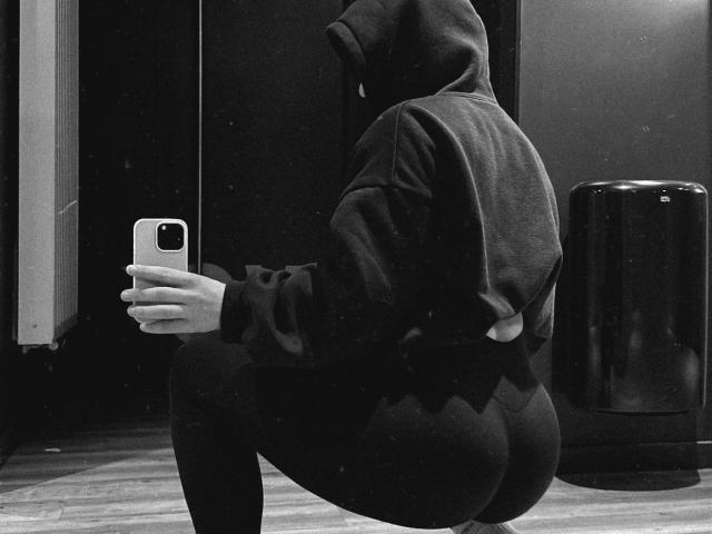MiraMint-hot, squatting, in black hoodie and tight pants, butt prominently, holding phone, black trash can in background, monochrome, textured, shadowy, holding phone with right hand.