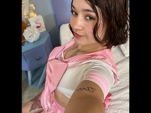 AnikaFleur with pink and white schoolgirl outfit, showing her tits, tattoos on arm and chest, dark wavy hair, red lipstick, sitting on white bed, blue nightstand in background.
