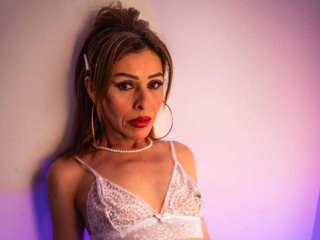 AngellCooper with brown hair, red lipstick, white lace top, pearl necklace, hoop earrings, standing against wall, looking seductively at camera, showing small tits, cute, sexy.
