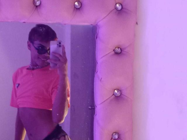 A mirror selfie of EstefaniRobles. EstefaniRobles has a slim build, short brown hair, and wears dark sunglasses. She's dressed in a neon pink crop top and black lace pants. She's holding a white smartphone in her right hand. The mirror is tufted with silver buttons, and the image has a purple tint. Her right arm is raised, revealing a flat stomach. The reflection shows her from the side.