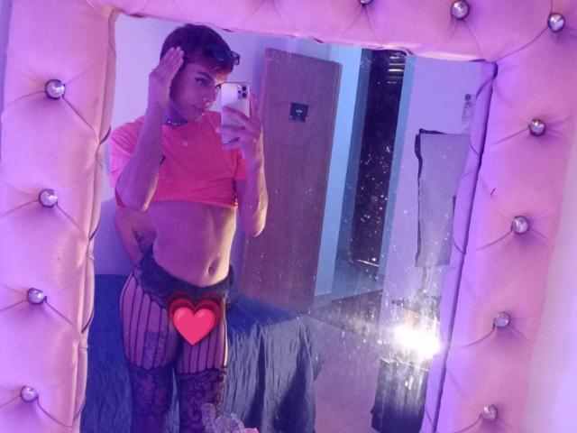 EstefaniRobles stands in front of a mirror wearing a pink crop top, black mesh pants with a heart over the crotch, and tattoos. She's holding a phone, looking at it.