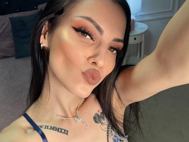 ScarletteStar with pouty lips, dark straight hair, heavy eyeliner, glowing skin, diamond earrings, necklace, tattoo on right shoulder, blue strappy top, raising her right arm, taking a selfie.