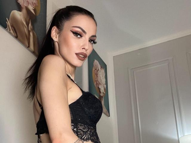 ScarletteStar with dark brown hair in a ponytail, wearing a black lace top, showing her tits, looking at the camera with dark makeup, in a white-walled room.