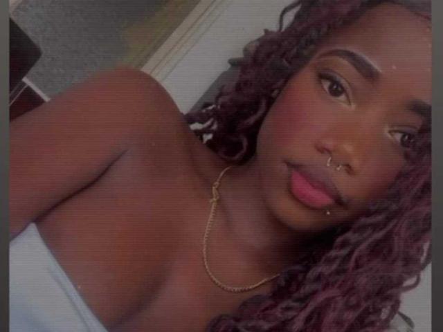 NyraSolenne has dark skin, pink lipstick, a nose ring, and a chain necklace, with curly purple-red hair, and is wearing a white strapless top, showing her bare shoulders.