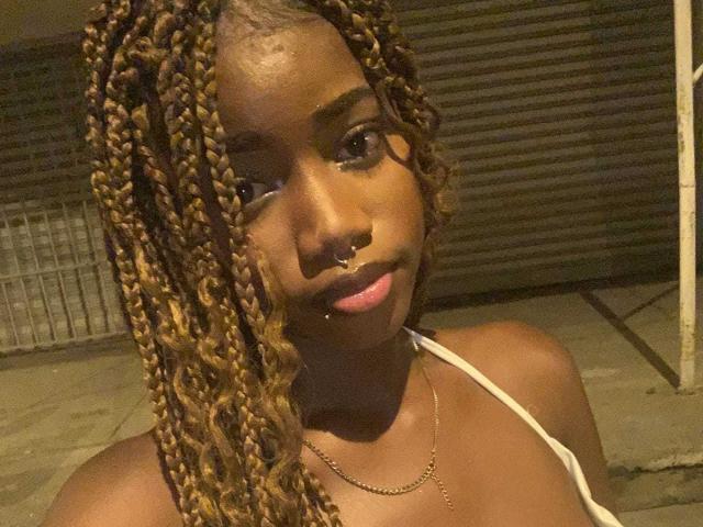 NyraSolenne has dark brown skin, curly blonde braids, a nose ring, and a lip piercing. She's wearing a white top with a gold necklace. Her tits are small.