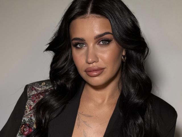 SheenaMelany with long black wavy hair, green eyes, glossy pink lips, black dress, and a tattoo on her chest. She's looking directly at the camera with a neutral expression.
