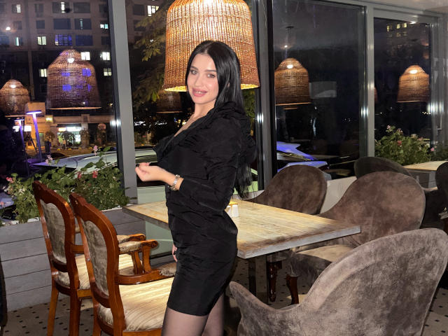 SheenaMelany's dark hair is visible above a large gray rectangle, with a warm, wicker lamp and a city night backdrop. Her face is mostly obscured.