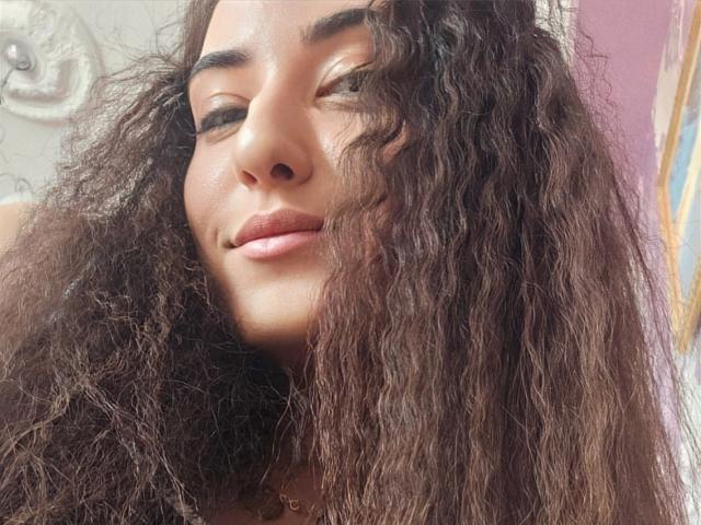 AlejandraDelaney's face with curly brown hair, slightly smiling, eyes half-open, subtle makeup, and a gold necklace, showing smooth skin and a cute, relaxed expression.