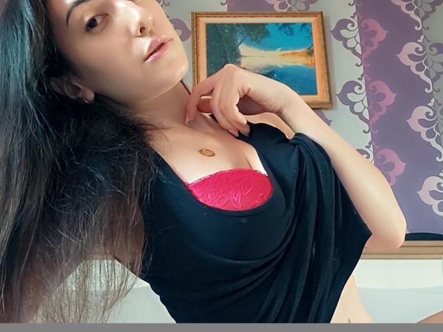 AlejandraDelaney with long black hair, black shirt, red lace bra, small gold necklace, and a confident smile, hand near chin, in a purple and patterned wallpapered room.
