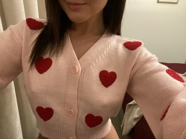 Dionisia wearing a pink cardigan with red heart embroidery, showing her tits, buttoned up, in a selfie. Her brown hair is down, lips slightly curved.