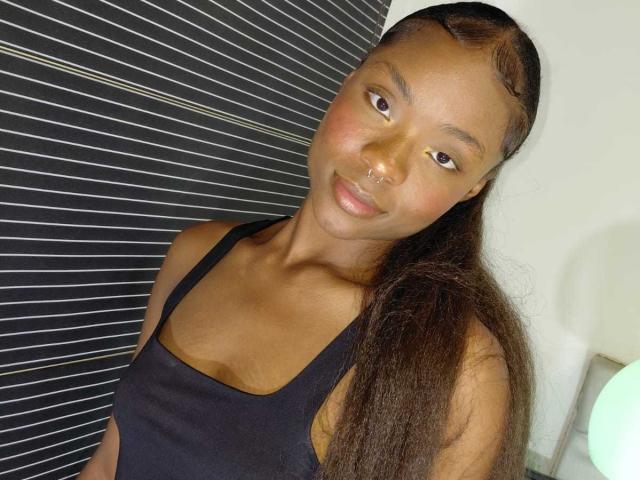Arianocthelle with long brown hair, nose ring, black tank top, small boobs, brown skin, subtle smile, looking at camera, soft light, slight tilt, relaxed pose.
