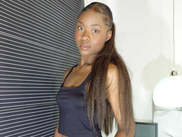 Arianocthelle with long, straight brown hair wearing a black tank top, showing small, perky tits, and a cute, subtle smile, standing in front of a striped wall.