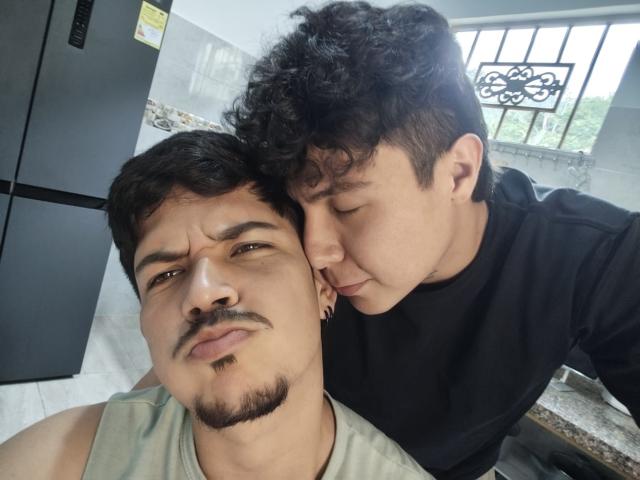 Danteelio with a mustache and goatee, wearing a green shirt, looking annoyed. Danteelio with curly hair, black shirt, kissing cheek. Kitchen background. Danteelio's face, Danteelio's kiss.