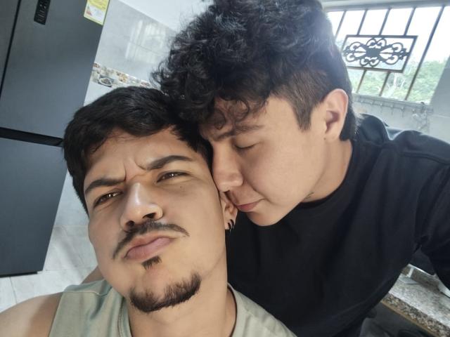 Danteelio with curly black hair, nose pressed to Danteelio's cheek, black shirt, looking cute, Danteelio with mustache, slightly pouty face, green shirt, kitchen background, daytime, sunlight.