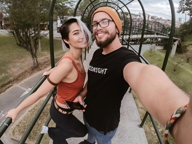 SpicyFran with short black-and-white hair, orange tank top, black arm band, and black pants, standing next to bearded SpicyFran in black shirt, orange beanie, taking selfie.
