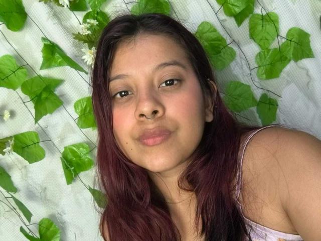 CoralineHale with dark brown hair, light brown skin, and subtle makeup, pouting with slightly parted lips, wearing a pink top, against a green leafy background.