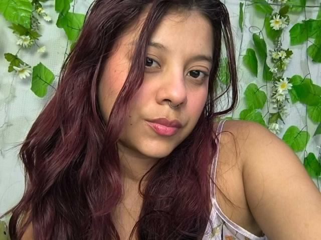 CoralineHale has medium brown skin, dark wavy hair with red highlights, a nose ring, and pink lipstick. Her small tits are partially visible in a white top.
