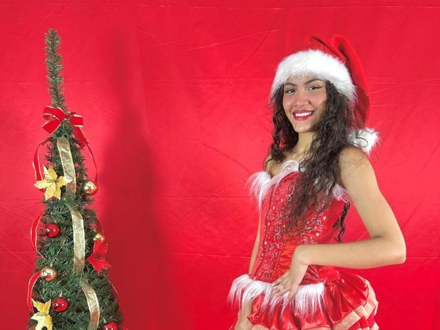 ValeryPoleth with long curly black hair, wearing a red Santa hat and red sequined corset with white trim, smiling, standing next to a small decorated Christmas tree.