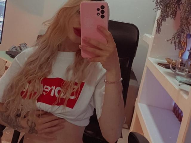 AlinaLee with long blonde hair, red lipstick, white shirt showing red text, holding pink phone, covering her tits, tattoo on right side, sitting in black chair, desk background.