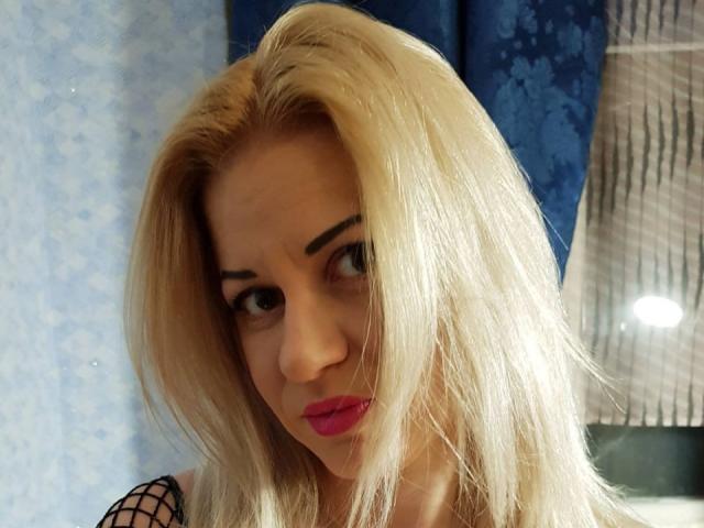 VladaVladaa with blonde hair, pink lipstick, and fishnet top, looking sideways with raised eyebrows. She has light skin, thick eyebrows, and a slight pout.