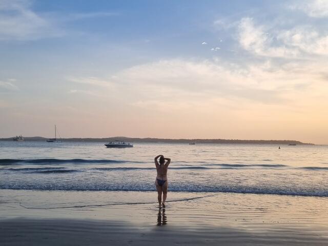 A beach scene with EstelaHot standing in shallow water, facing the ocean. She's topless, wearing blue bikini bottoms, with hands up, holding hair. The water reflects her silhouette. Waves gently roll in. A boat is in the mid-ground, and distant land is on the horizon. Sky is blue with light, wispy clouds. EstelaHot's reflection is clear in the water. Slang: She's chillin' in the water, lookin' all relaxed.