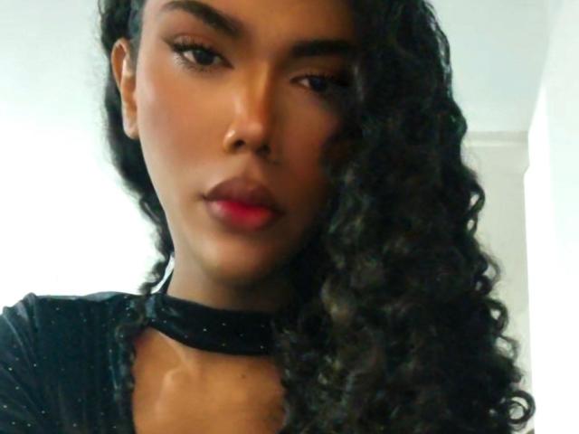 NinfTrans with dark curly hair, black choker, and red lips, looking serious, has smooth brown skin, and is wearing a black top. Boobs not visible. Pussy not visible.
