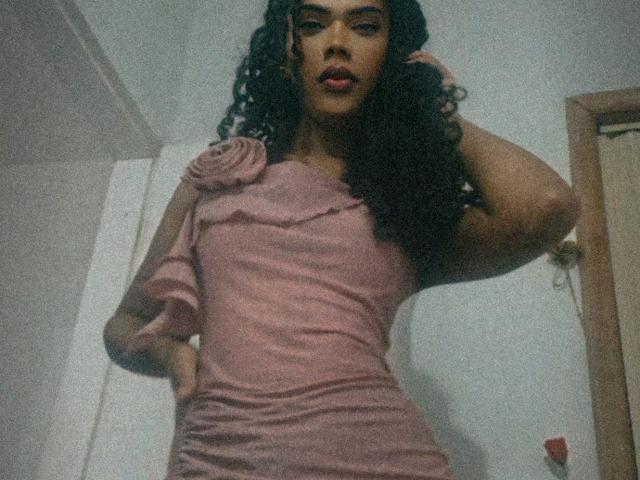 NinfTrans in a pink dress, standing with one hand on her hip, the other hand in her curly black hair, looking down at the camera with red lipstick.