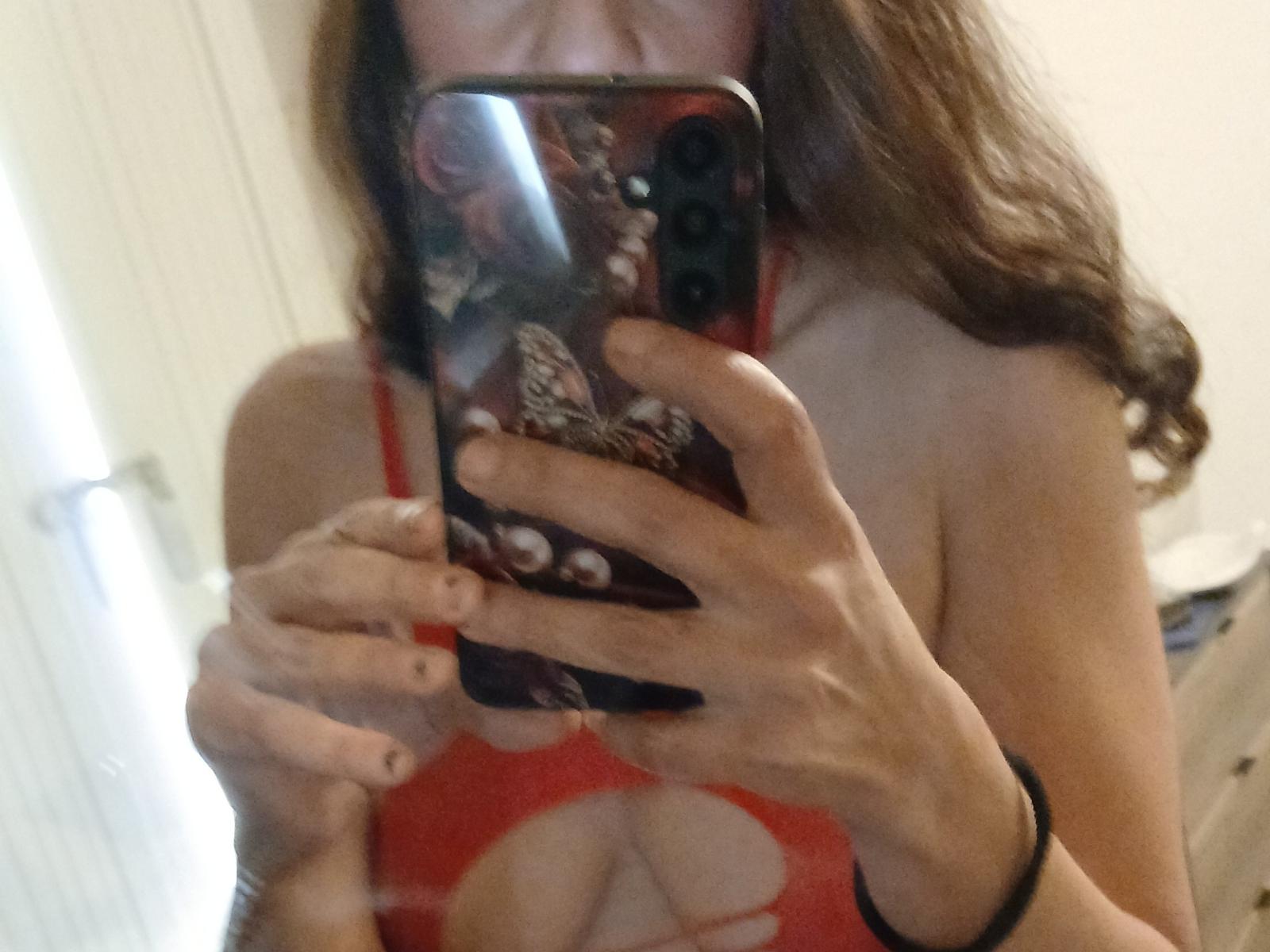 Maitressebdsm06 tira uma selfie no espelho com o celular, cobrindo o rosto. Ela está vestindo uma blusa vermelha com recortes, que mostra seus seios, e tem cabelos longos castanhos..