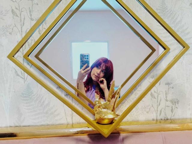 Mirror reflection of LolyYunno with red hair, holding a smartphone in a diamond-shaped frame. LolyYunno has a relaxed, slightly playful expression, with one hand touching her face. The mirror has a gold geometric frame with a golden ornamental base. LolyYunno is wearing a purple top. The background shows a light-colored wall with subtle floral patterns. The reflection is centered in the diamond frame, with the smartphone's patterned case visible. The overall composition is balanced, with the gold frame adding a touch of elegance. The mirror's reflection is sharp and clear.