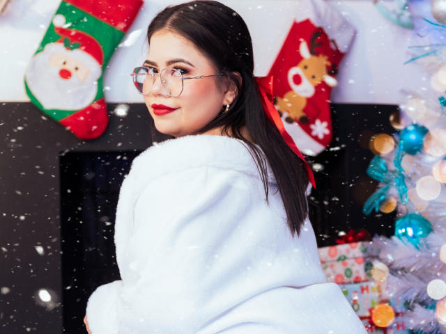 LilyRousee with long dark hair, wearing glasses and a white fluffy coat, looks over her shoulder. She has red lipstick and a slight smile. Snowflakes fall around her. Background includes Christmas stockings with Santa and reindeer designs, a blurred Christmas tree with blue ornaments, and wrapped gifts. The image uses bright colors and a soft focus on the background, highlighting LilyRousee as the central figure. Slight bokeh effect adds to the festive atmosphere.