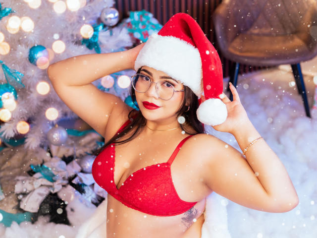LilyRousee with red bra, Santa hat, glasses, red lipstick, and earrings, showing her tits, hands in hat, snowflakes, and white Christmas tree with lights in background.