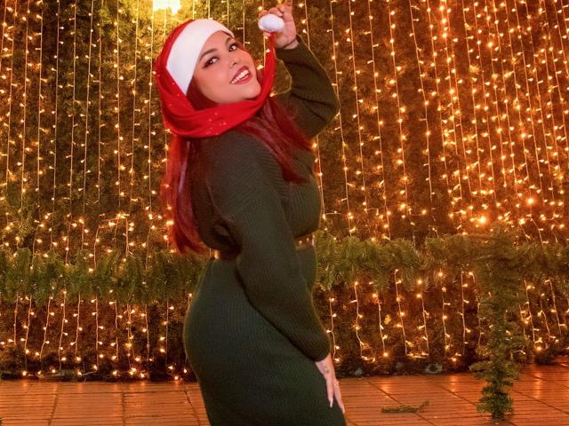 MorganMoretz with red Santa hat, green sweater dress, holding Santa hat, smiling, showing curvy ass, long red hair, standing in front of string lights.