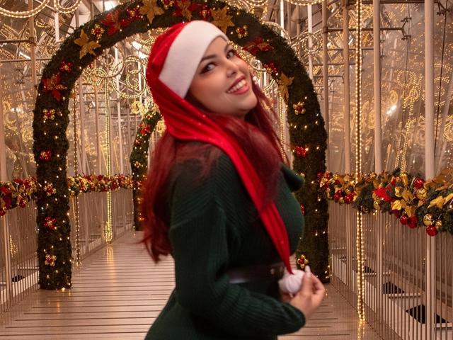 MorganMoretz with red hair, green sweater, and Santa hat, smiling at the camera, holding Santa hat with white pom, showing her ass slightly in the background.