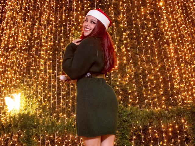MorganMoretz with red hair, wearing a Santa hat, green sweater, and high-waisted skirt, smiling at camera, showing her ass, standing in front of glowing string lights.