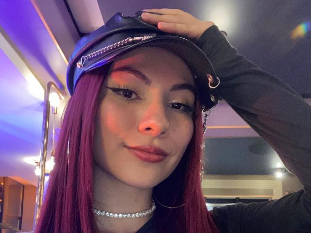 ValeryHarllow with red hair, wearing a black leather cap, black long-sleeve shirt, and choker, has a nose piercing and smirks, hand in cap, in a dimly lit room.
