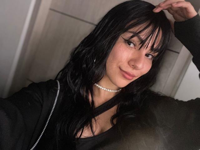 ValeryHarllow, avec de longs cheveux noirs et une frange, portant un haut noir, un collier de perles, souriante, la main droite dans les cheveux, yeux marrons, lèvres roses claires, poitrine moyenne, peau claire..
