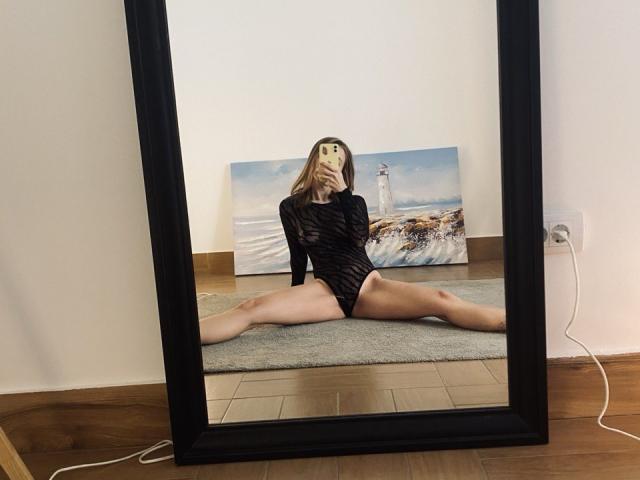 Nympheexe is in a black, sheer, long-sleeve bodysuit, sitting on a carpet, spreading her legs wide in a mirror reflection, showing her pussy and shaved ass.