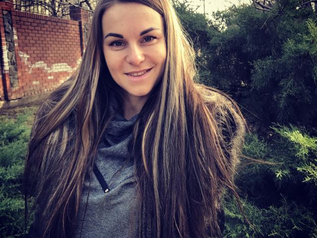 BelleLisaG has long brown hair, a gray hoodie, and a slight smile. She's standing in front of greenery with a brick wall in the background.