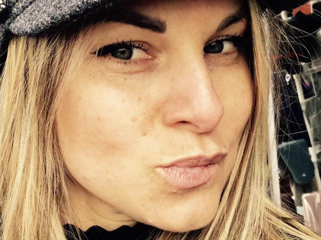 Close-up of BelleLisaG's face with blonde hair, green eyes, and slightly pouty lips. She wears a grey flat cap and has light skin with small freckles. Her eyebrows are dark and slightly raised. The image focuses on her face, with subtle texture in her skin and a natural, slightly messy look. The background is blurred, making BelleLisaG's face the main focus. The composition is tight, emphasizing her facial features.