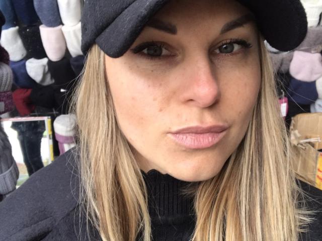 BelleLisaG with blonde hair, black turtleneck, and black hat, looking at camera with slight smirk, visible freckles, and long eyelashes. Boobs not visible.