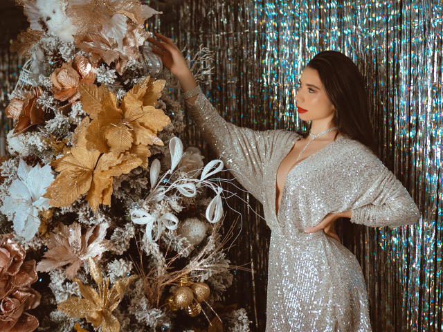 Bellasweeth, mit ihren langen, dunklen Haaren, trägt ein glitzerndes, silbernes Kleid und berührt eine goldene und weiße Blumenanordnung. Ihre roten Lippen bilden einen Kontrast zu ihrer blassen Haut..