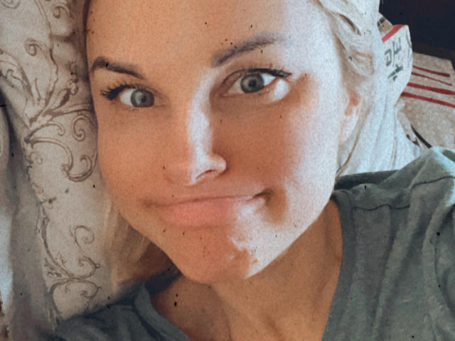 Close-up of BelleLisaG lying down, face slightly tilted. She has light blonde hair, green eyes, and a subtle smile. She wears a green shirt, with visible freckles on her face and a small white substance on her chin. The background has a patterned pillow with swirling designs and red stripes. The image has a grainy texture, adding a rough, almost vintage feel. Her makeup includes long, dark eyelashes. Her skin is light with a slightly pinkish hue.